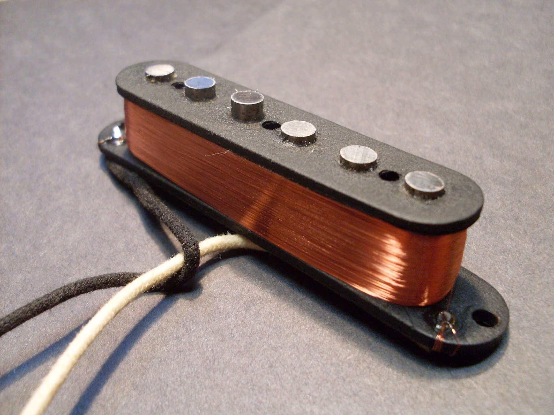 A single-coil pickup off the guitar: six cylindrical alnico magnets standing through black flatwork, with thousands of turns of fine enamelled copper wire wound around them, and two cloth-covered leads.
