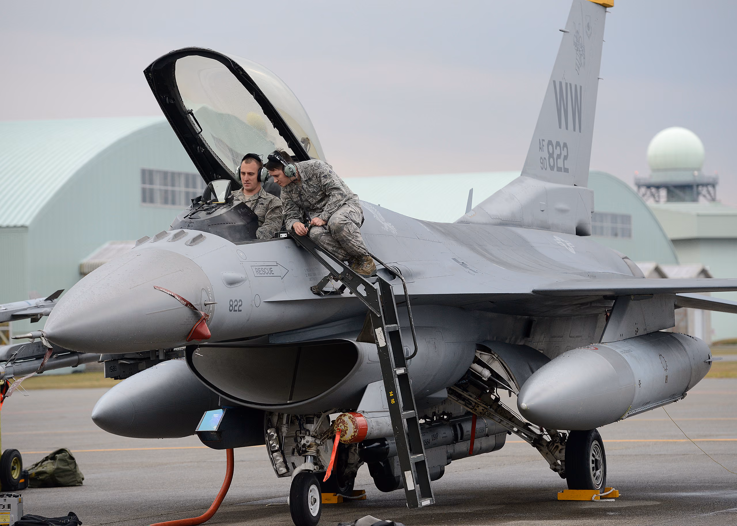 Two USAF avionics technicians in front of an open F-16 fuselage panel, running a systems check with test equipment plugged into the aircraft