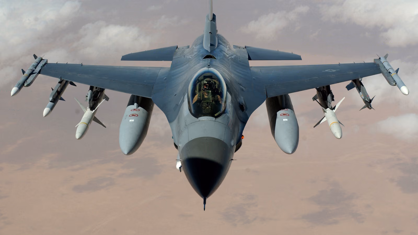 An F-16 seen head-on in level flight against a dust-coloured sky, wingtip missiles and underwing stores visible, the pilot's helmet just showing under the canopy