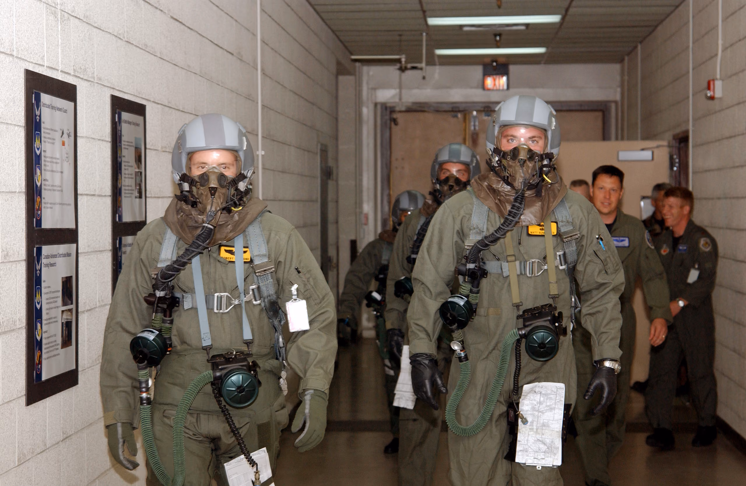 Two F-16 instructor pilots in flight suits walking side by side toward a simulator research facility, helmets and gear in hand