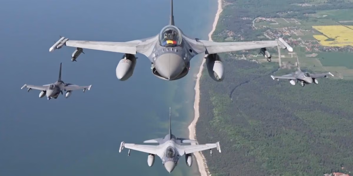 Four F-16s in loose formation over a coastline, seen from above and behind, farmland and forest inland of a long sand beach