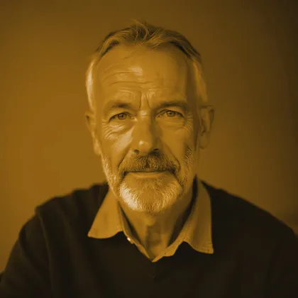 A staff portrait, toned in the site's gold and bronze. An older man with grey hair and a grey beard, in a dark sweater over an open collar, facing the camera square on with an even, unhurried expression against a plain warm wall.