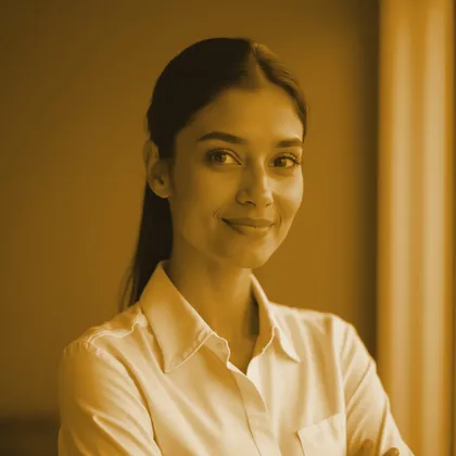 A staff portrait, toned in the site's gold and bronze. A younger woman with dark hair tied back, in a white collared shirt with her arms folded, smiling at the camera against a warm wall crossed by a soft band of window light.
