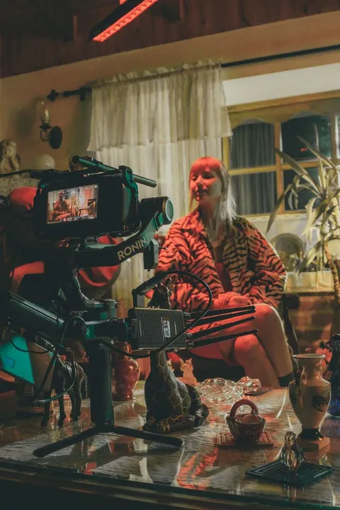 A small film crew operating a camera on a tripod alongside a lighting rig inside a cozy, real indoor film set.