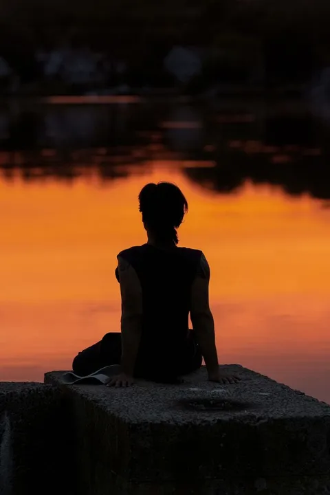 A silhouetted figure sitting quietly by a calm lake at sunrise, alone, no camera crew or screen in sight.