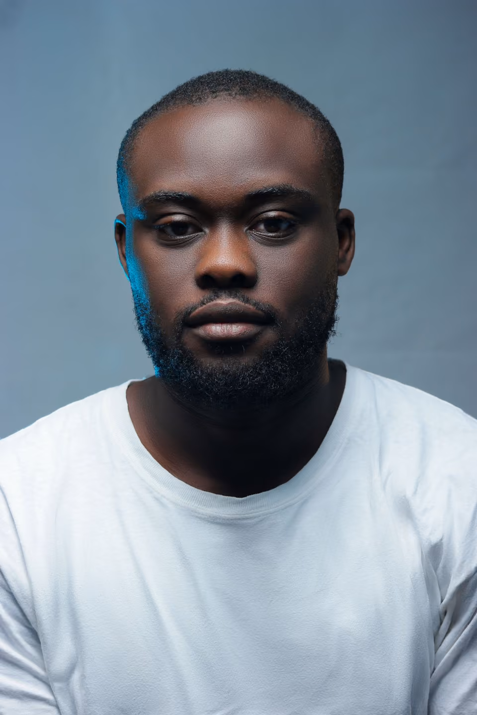 A studio headshot of a man with a short beard, looking directly at the camera against a plain grey backdrop, wearing a plain white shirt.