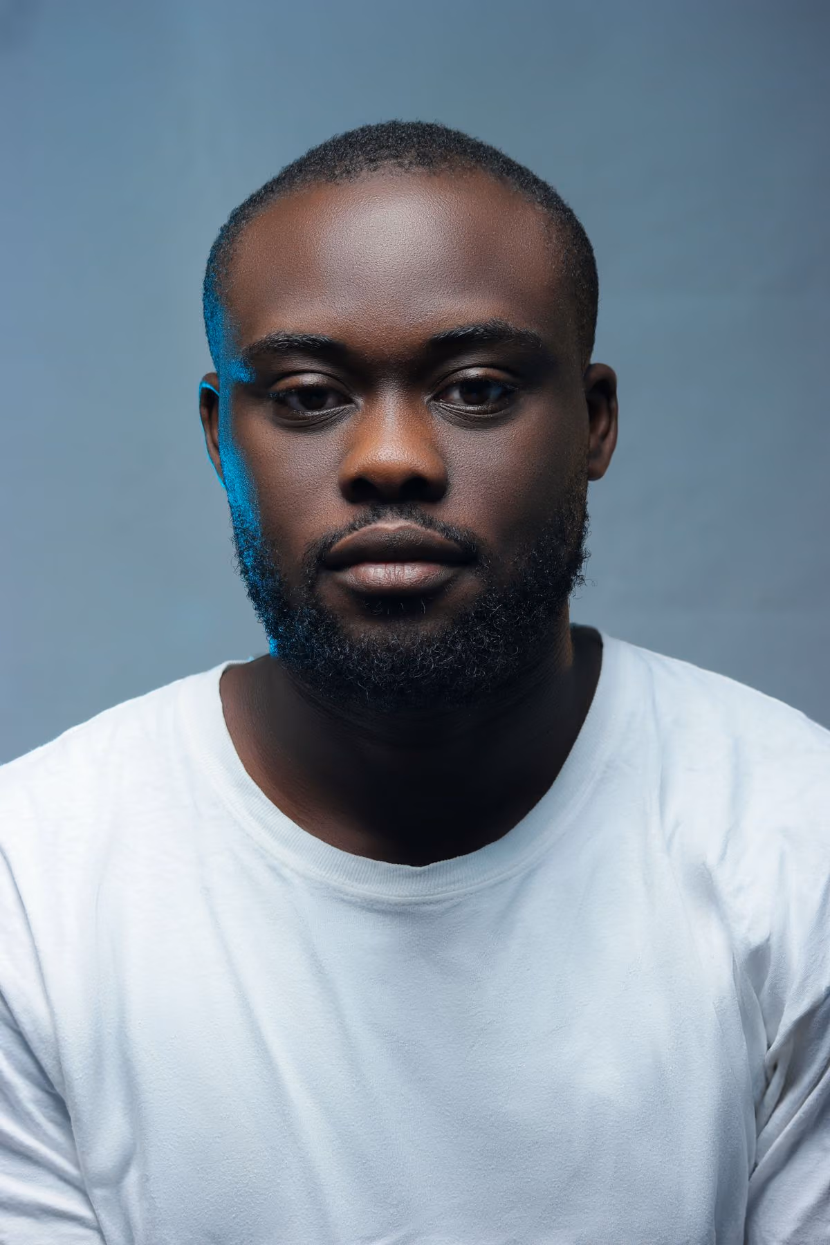 A studio headshot of a man with a short beard, looking directly at the camera against a plain grey backdrop, wearing a plain white shirt.