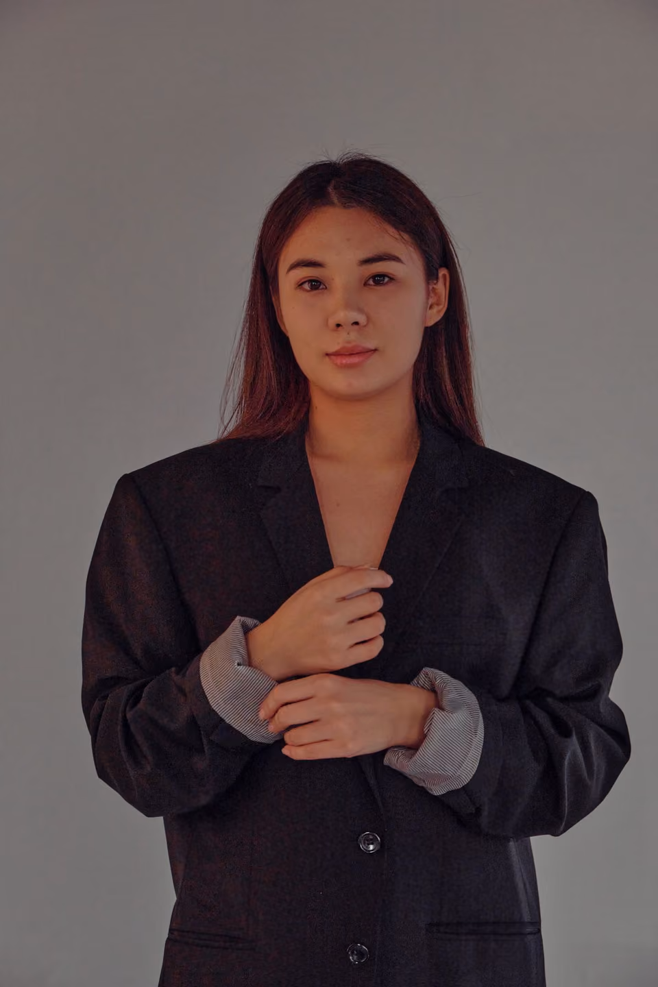 A studio portrait of a woman with long dark hair in an oversized black blazer, hands clasped at the lapel, looking directly at the camera against a plain grey backdrop.