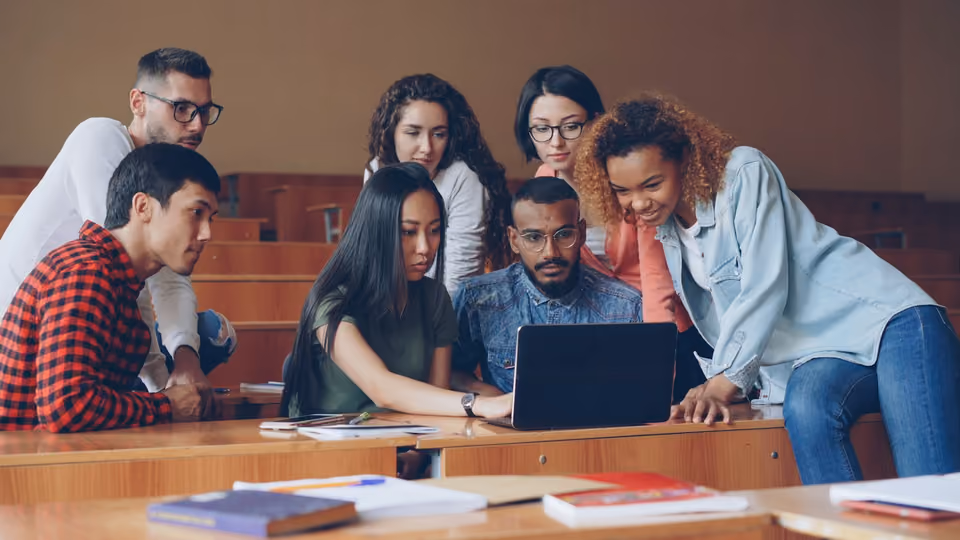 Seven students crowded around a single open laptop on a lecture-hall desk, all looking intently at the screen