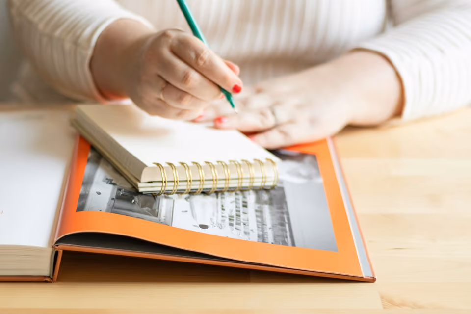 A hand with red nail polish writes with a pen in a spiral notebook resting atop an open photo album showing a black-and-white photograph