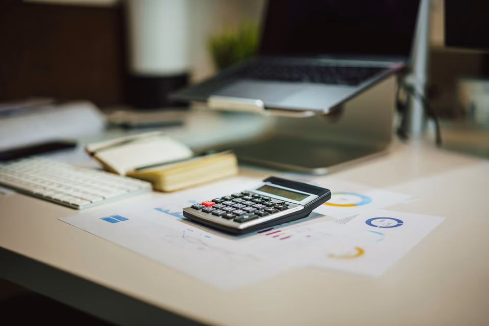 A calculator sitting on top of printed financial charts and graphs on a desk, with a laptop and keyboard blurred in the background