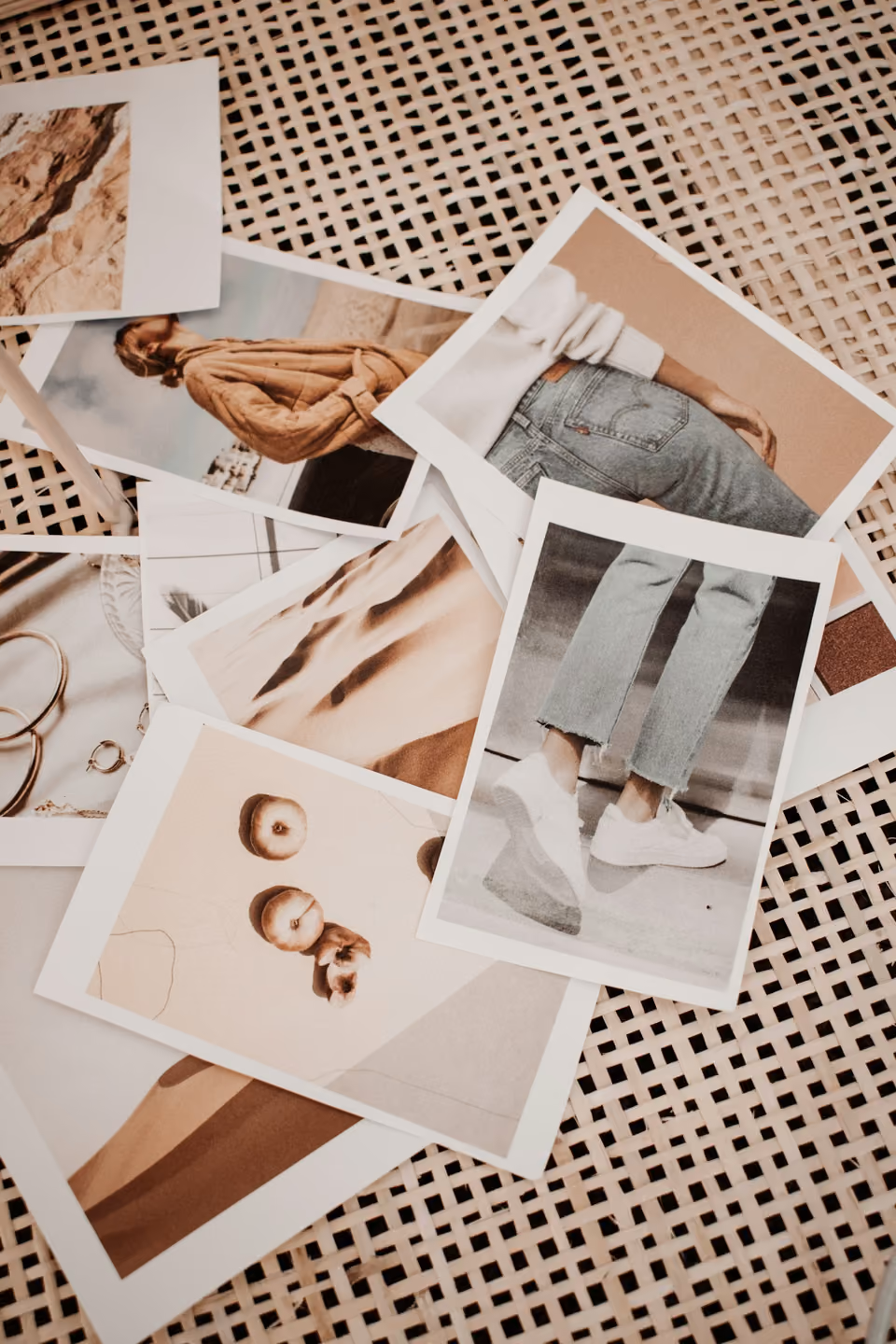 A scattered pile of printed photographs — fashion shots, a landscape, jewellery — spread across a woven rattan surface for comparison