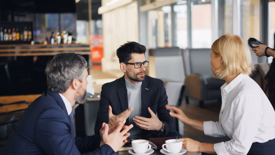 Three people in business attire gesturing and talking animatedly around a small table with coffee cups