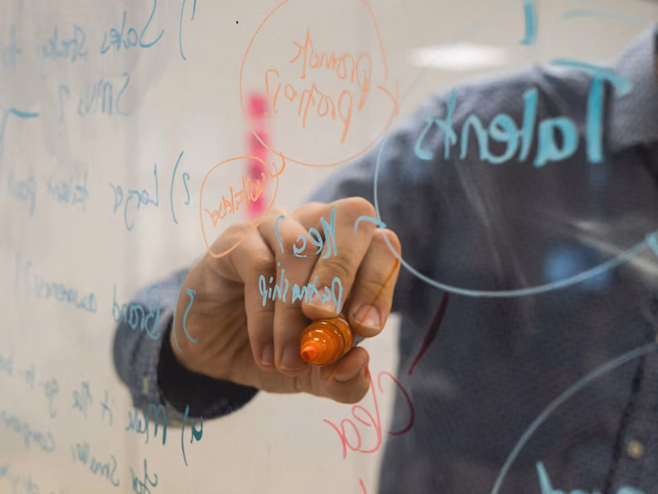 A hand draws circled labels and connecting arrow lines with a marker on a glass whiteboard covered in diagram notes