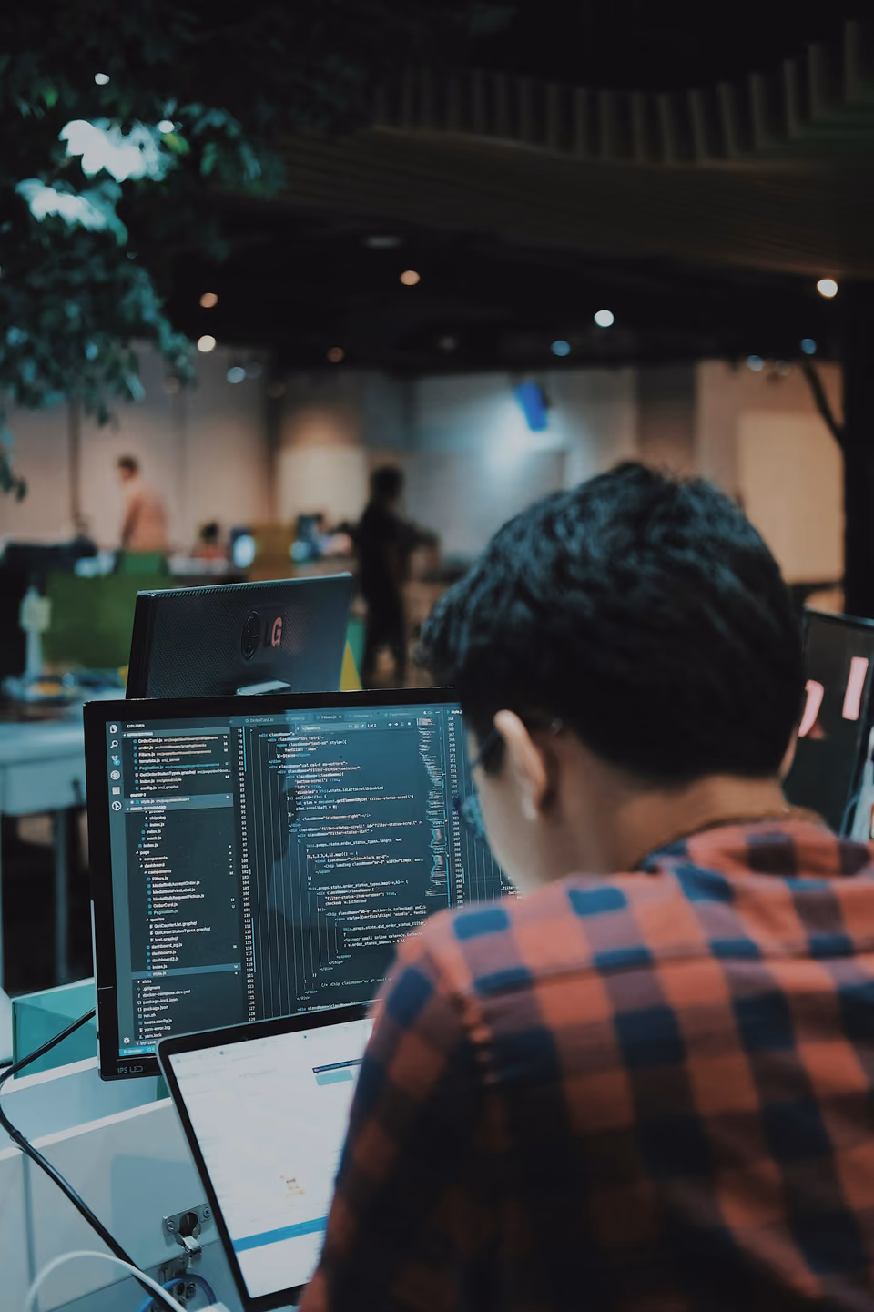 A person viewed from behind, working at dual monitors showing a code editor with a file tree and source code open