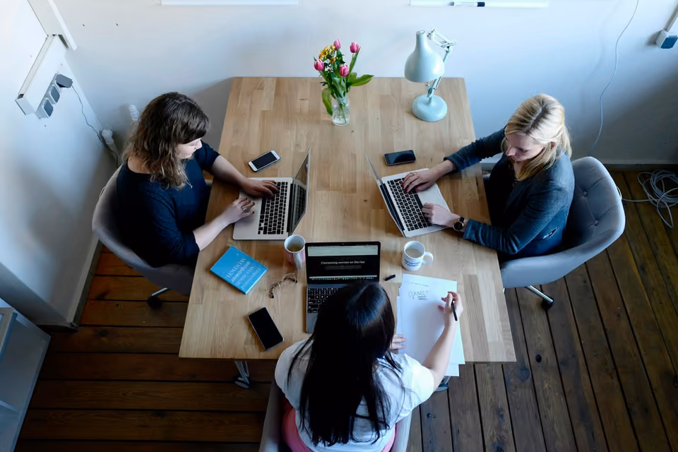 Overhead view of three women at a table with laptops open, one writing on a sheet of paper