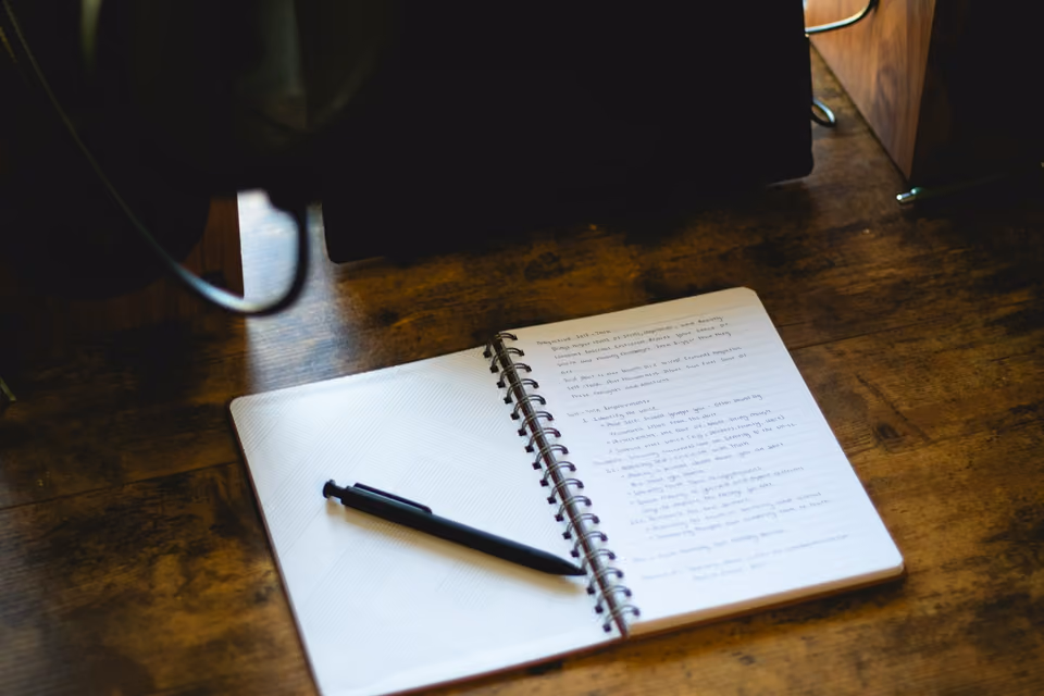 An open spiral notebook filled with handwritten reflective notes, a pen resting across the page, on a dark wooden desk
