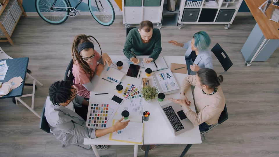 Overhead view of five colleagues around a table covered in colour swatches, charts, coffee cups and a laptop, in a creative studio