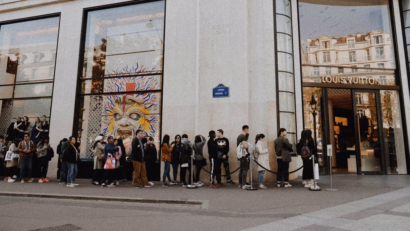A queue running along the pavement outside a shop front, bunched in places and thin in others
