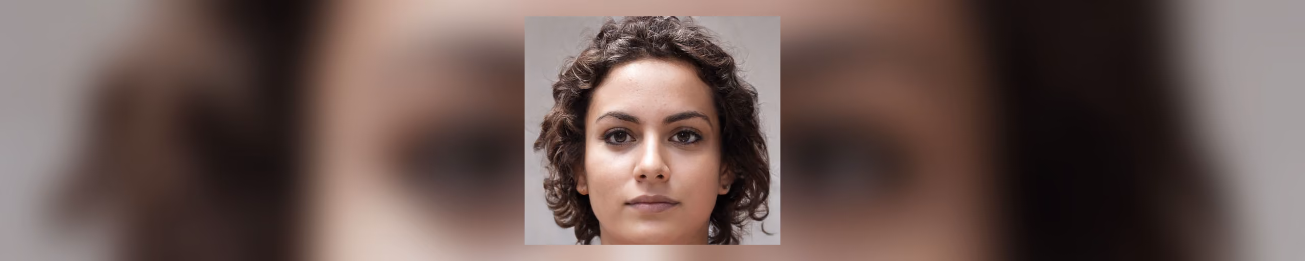A photographic portrait of a woman with short dark curly hair, a neutral expression, facing the viewer against a plain grey background.