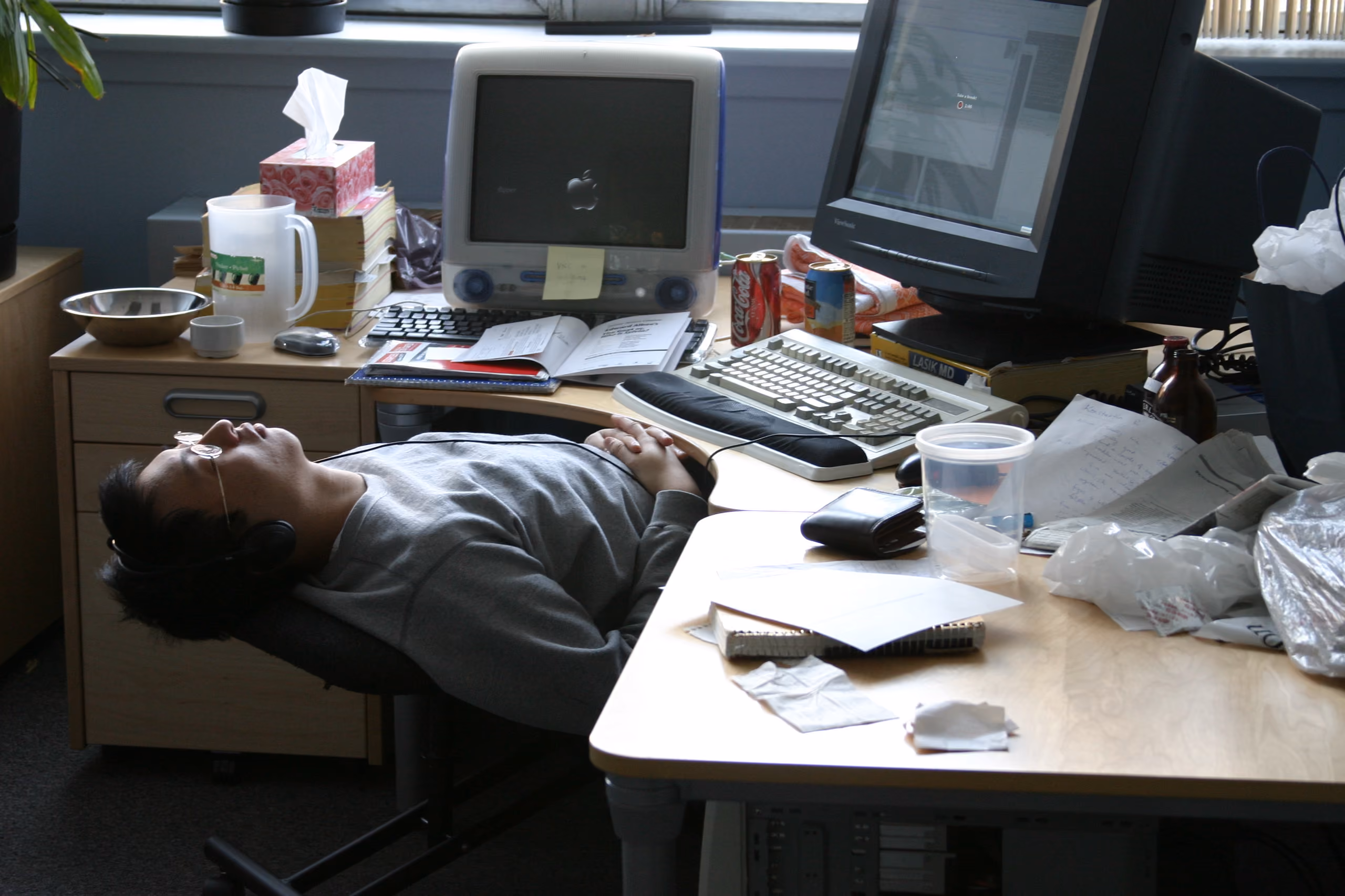 A person reclining with headphones on beside a cluttered office desk — an old monitor, a tissue box, a mug, scattered papers — eyes closed, mid-shift