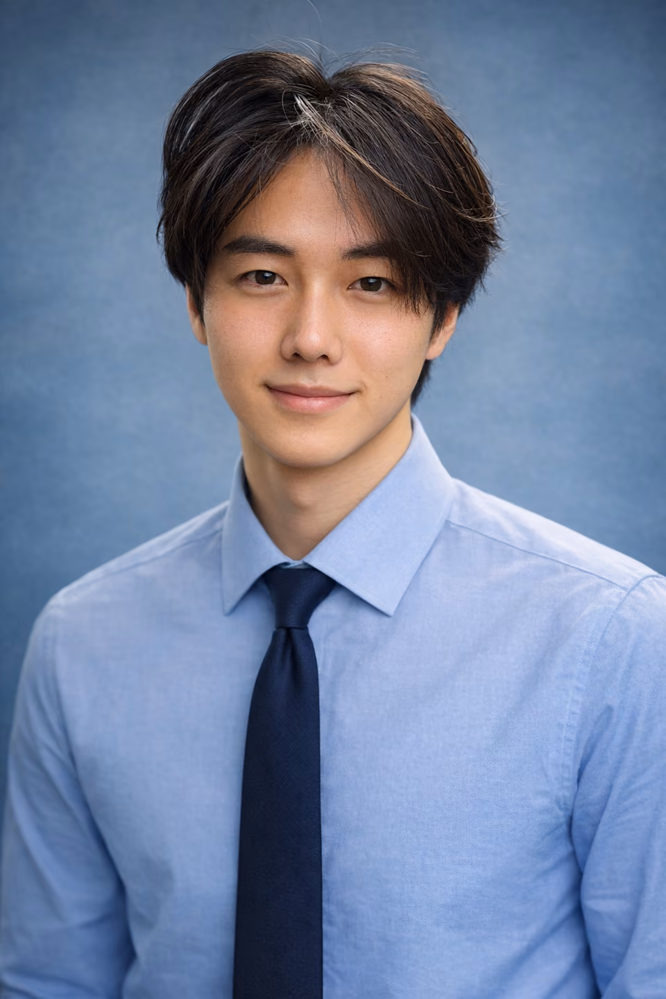 A studio headshot of a young man with dark hair, smiling slightly, in a light blue collared shirt and navy tie against a blue backdrop