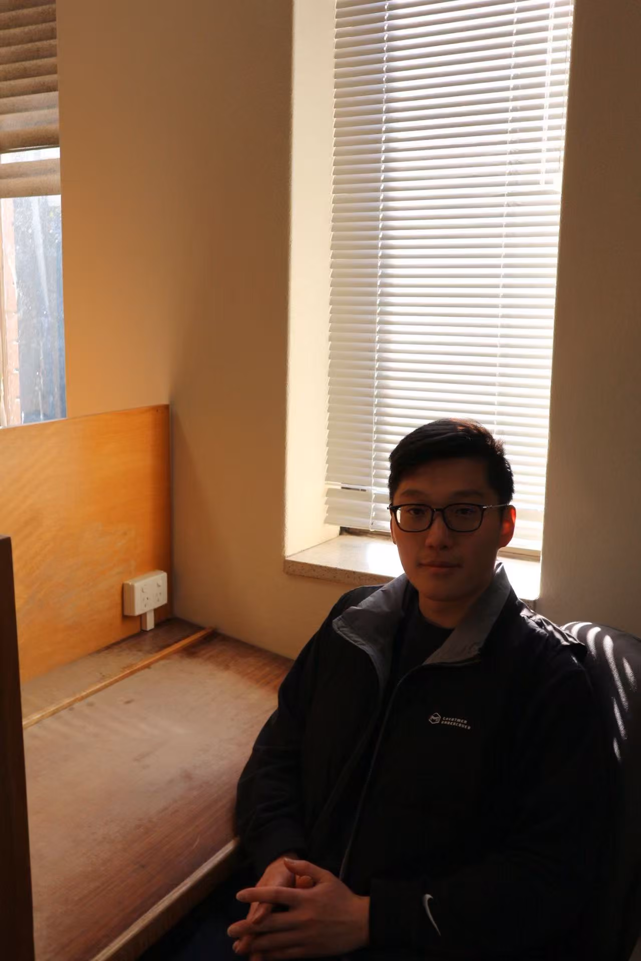 A young man with glasses and dark hair, wearing a dark jacket, seated in front of a sunlit window with blinds