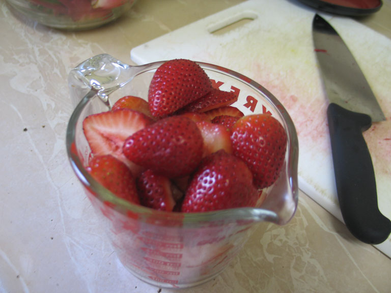 A white plastic cutting board and a kitchen knife on a countertop, with strawberries being sliced in the foreground