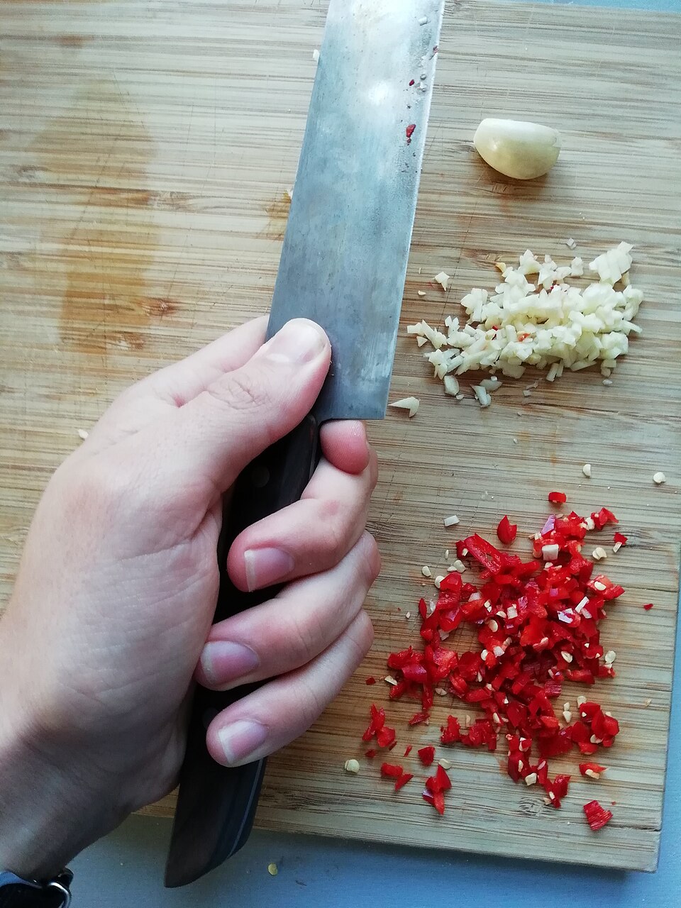 Overhead view of a hand gripping a chef's knife in a pinch grip, thumb and forefinger ahead of the bolster against the flat of the blade, cutting garlic and chilli on a board