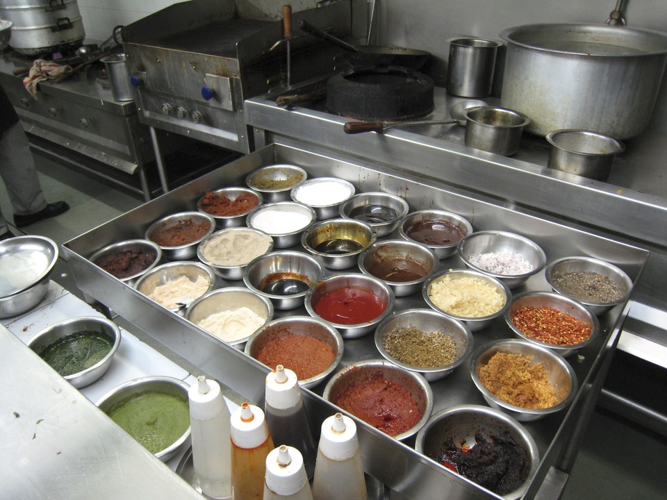 A commercial kitchen prep station with rows of stainless steel bowls holding sauces, spices and pastes staged ready for service