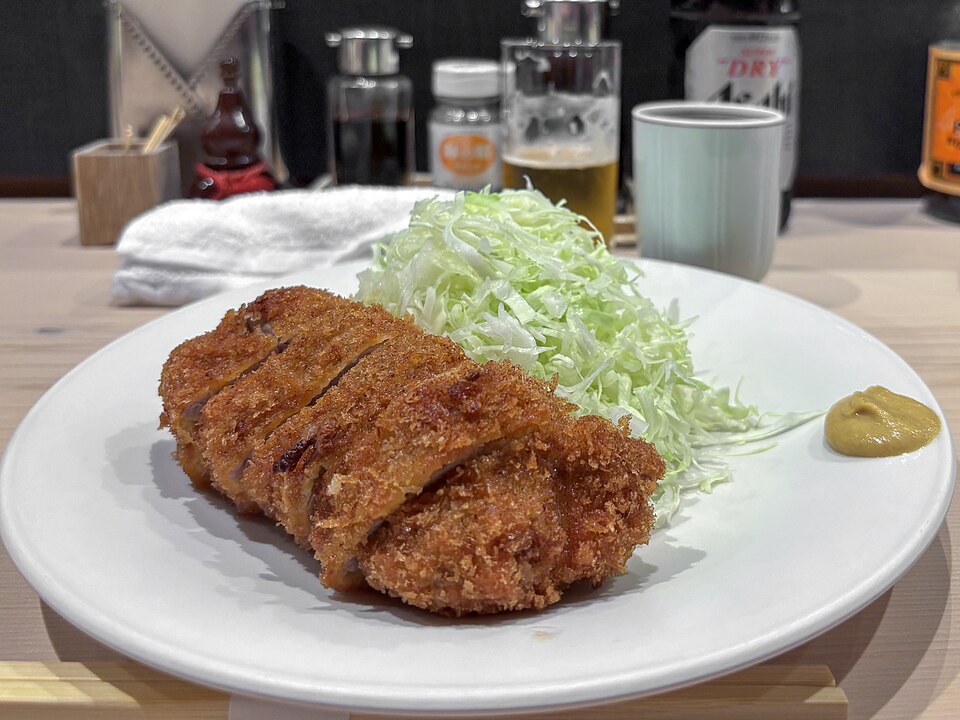 A sliced breaded pork cutlet on a white plate beside a mound of shredded cabbage and a dab of mustard, on a wooden counter with condiment bottles and a glass of beer blurred in the background.