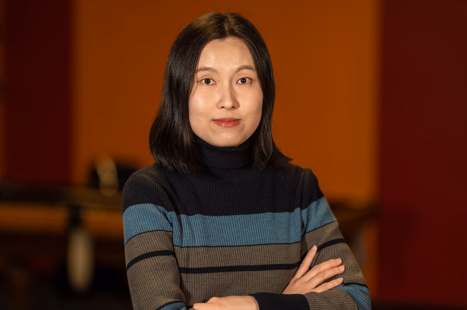May Rhee, arms folded, looking directly at the camera against a warm orange wall