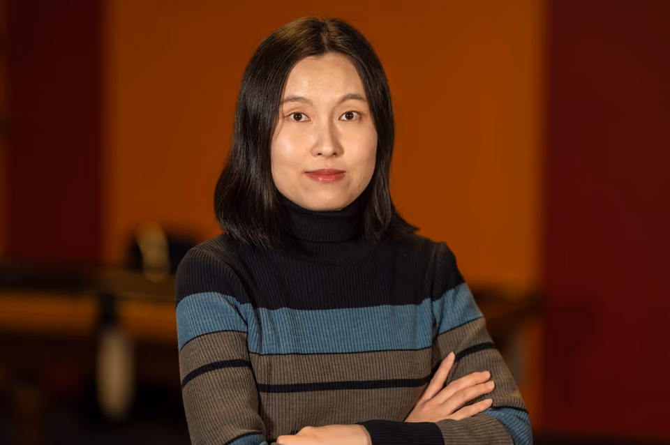 May Rhee, arms folded, looking directly at the camera against a warm orange wall
