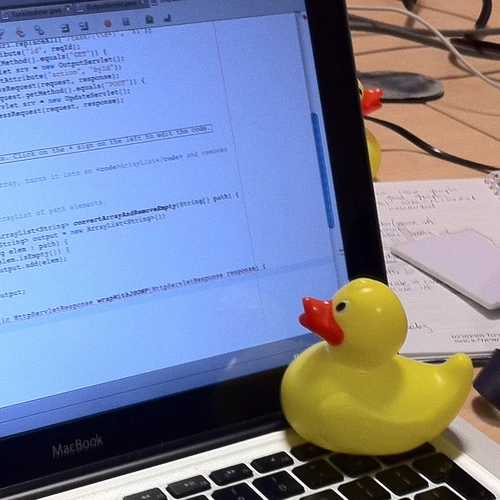A yellow rubber duck sits on a MacBook keyboard beside a screen of Java code.