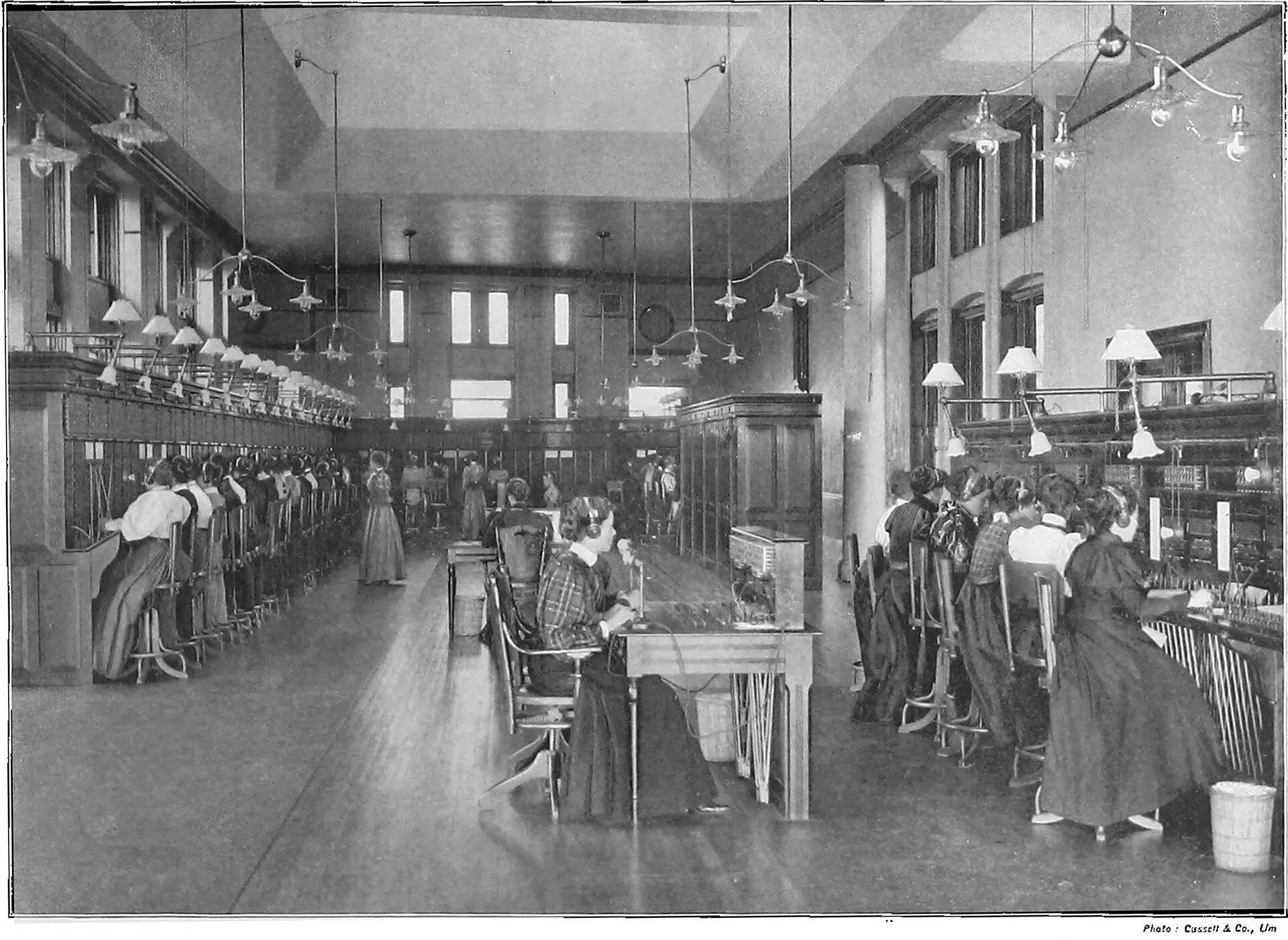 A long hall in a Montreal telephone exchange in the late 1890s. Two facing banks of switchboards run the length of the room, with about twenty operators at each, headsets on, reaching up into the jackfields. One supervisor sits at a desk in the aisle between them.