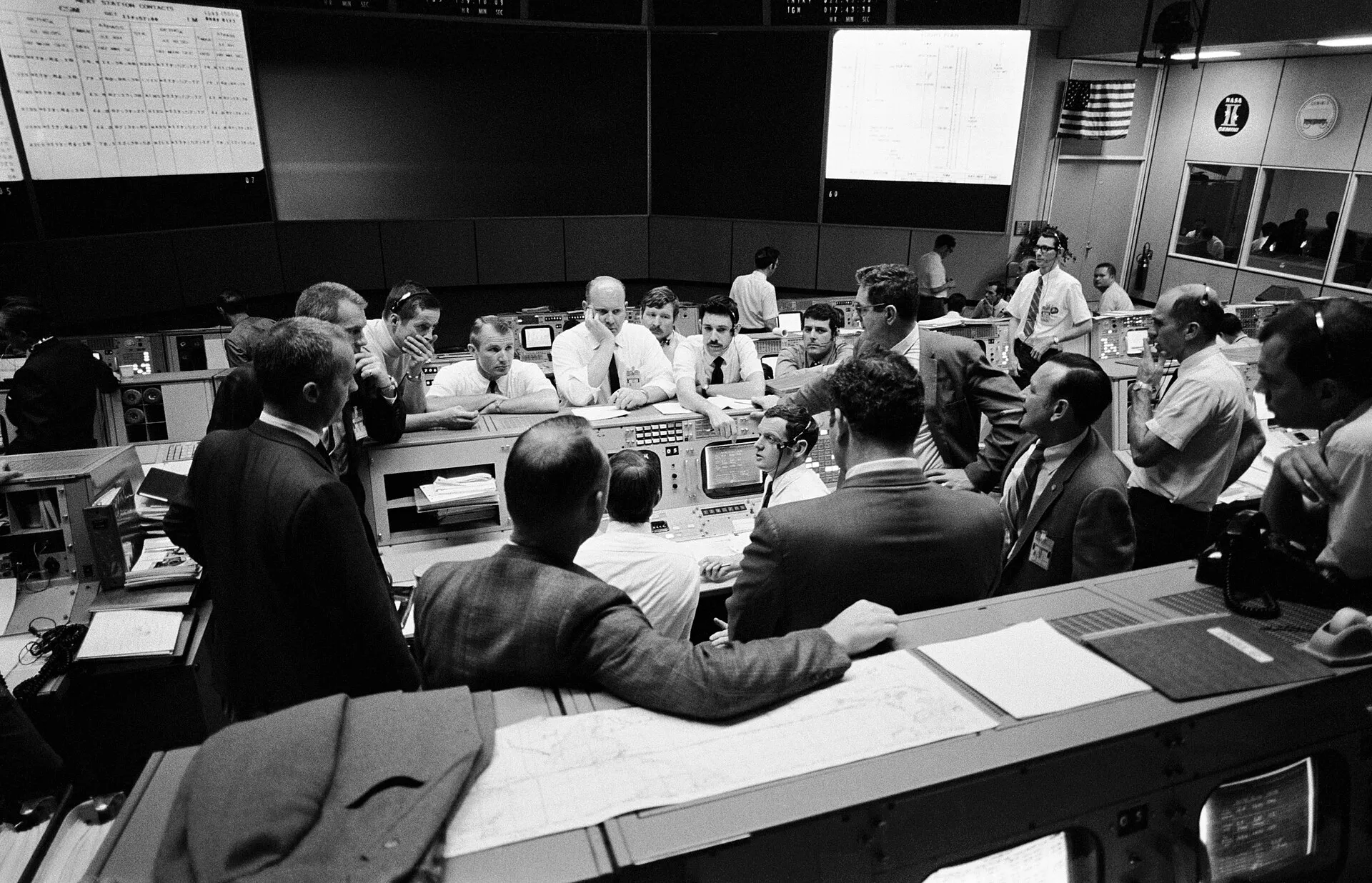 Mission Control in Houston during Apollo 13's return, April 1970. Two dozen controllers and managers crowd around a console, several leaning in, one with a hand over his mouth. Flight plans and open documents cover every surface.