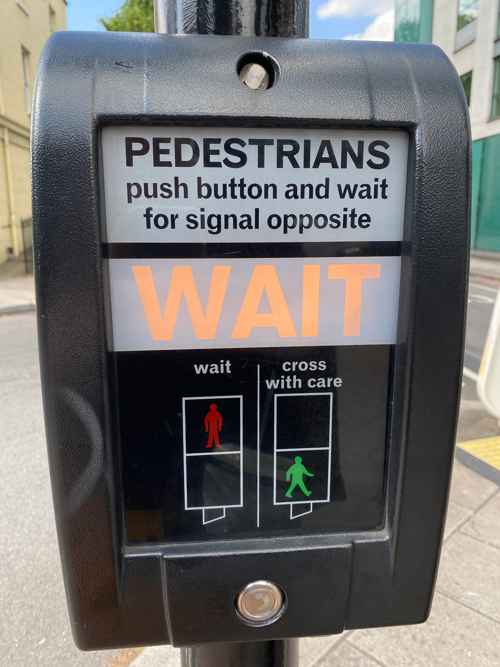 A pedestrian crossing button with an illuminated orange WAIT indicator. The instructions read: “PEDESTRIANS push button and wait for signal opposite”, with red wait and green cross-with-care symbols.